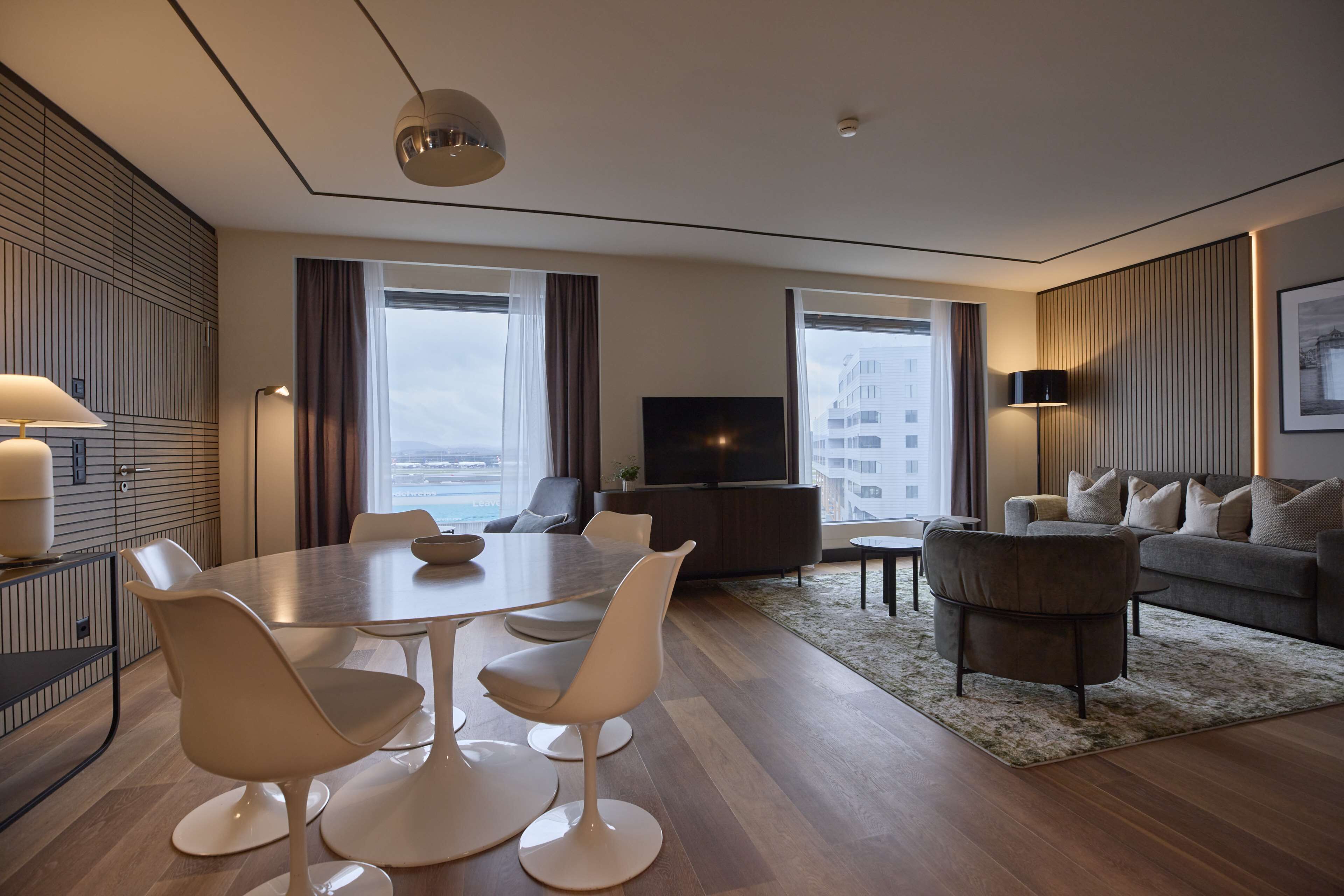 A modern hotel room with a dining area, a sofa set, a TV, and large windows offering a city view.