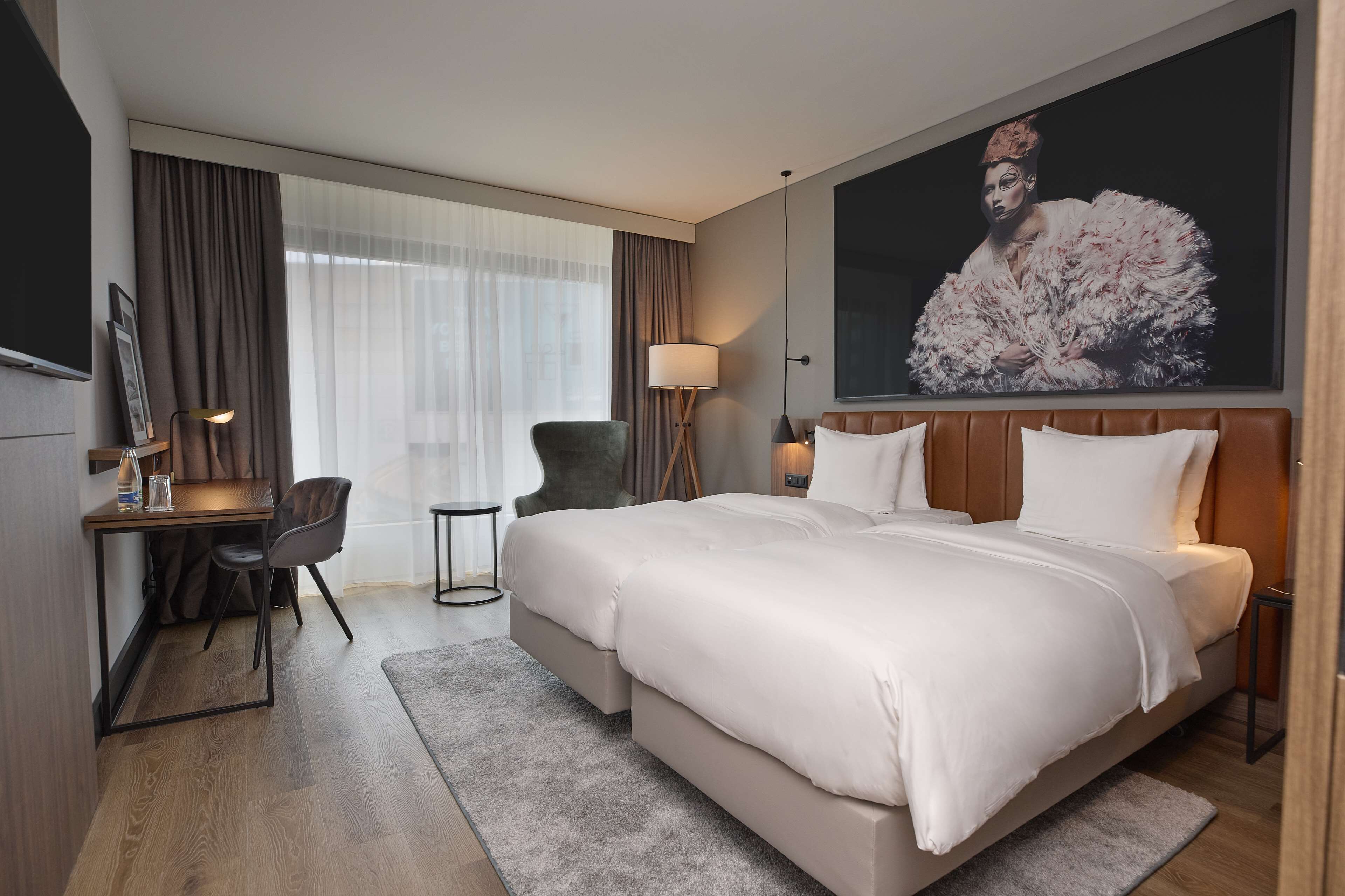 Een hotelkamer met een groot bed, een bureau met stoel, een klein tafeltje en een groot raam met gordijnen.