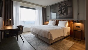 Habitación de hotel moderna con una cama grande, un escritorio y una silla. Cuenta con un amplio ventanal con cortinas, un cuadro en la pared y una mesita de noche con una lámpara.