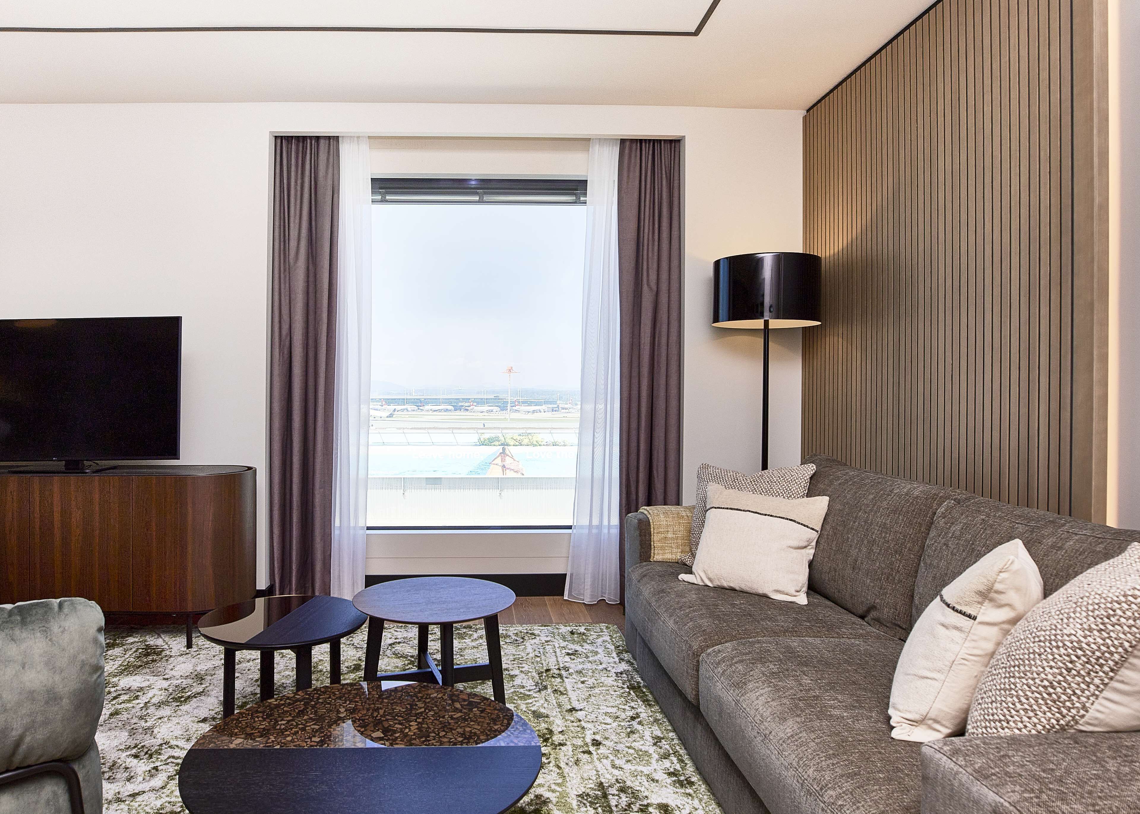 Una habitación de hotel moderna con un ventanal grande, un televisor de pantalla plana, un sofá, una mesita de café y una lámpara de pie.