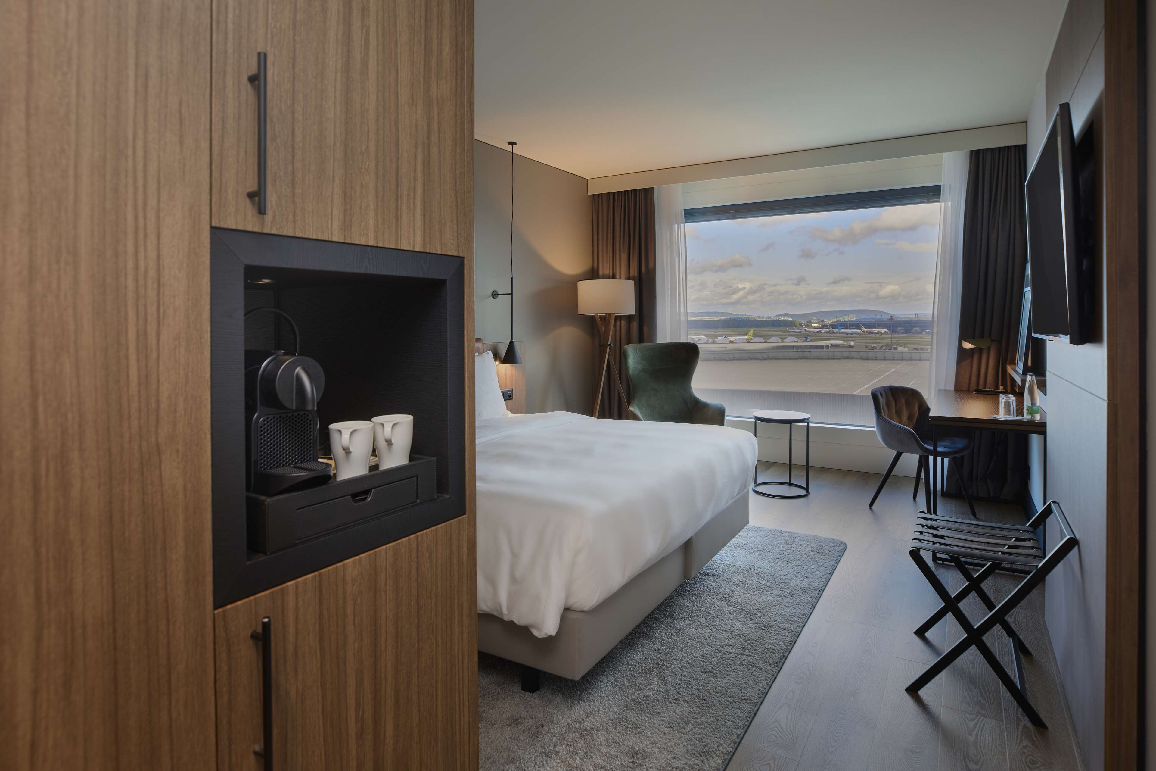 Hotelkamer met een bed, een bureau, een stoel, een televisie en uitzicht op het vliegveld.