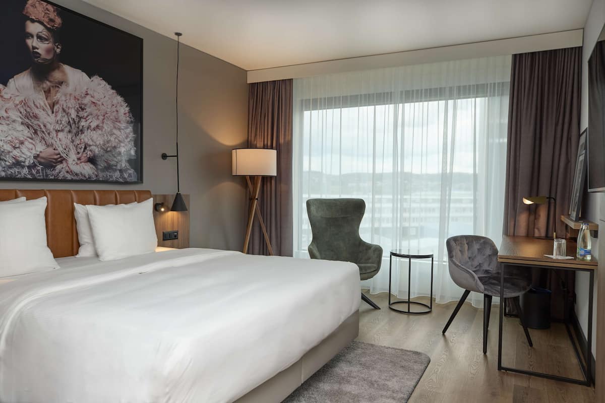 Habitación de hotel con una cama grande, un escritorio, una silla, una lámpara y vistas a la ciudad.