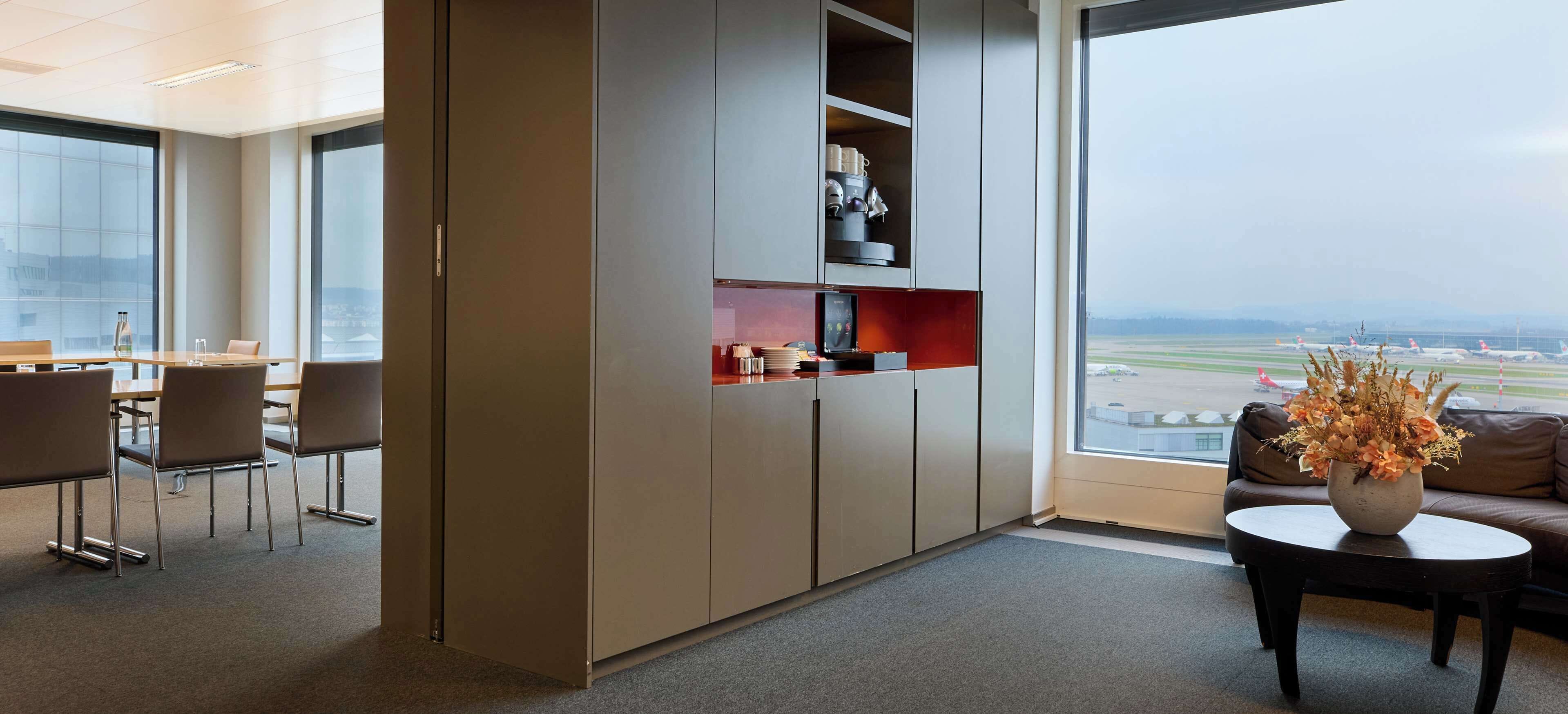 A modern office space with a large window overlooking an airport. It includes a seating area with chairs and a table, a kitchenette with cabinets, and a decorative vase on a coffee table.