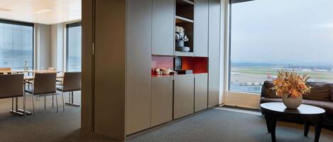 Un espacio de oficina moderno con un amplio ventanal que da a un aeropuerto. Incluye una zona de estar con sillas y una mesa, una kitchenette con armarios y un jarrón decorativo sobre una mesa baja.