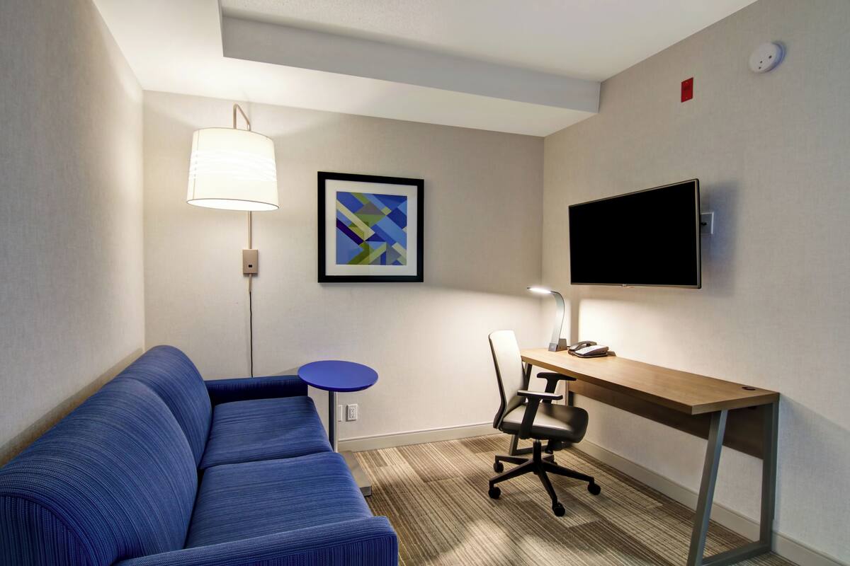 Une chambre d’hôtel avec un canapé bleu, une petite table ronde, un bureau avec une chaise, un téléviseur fixé au mur et une œuvre d’art abstraite encadrée accrochée au mur.
