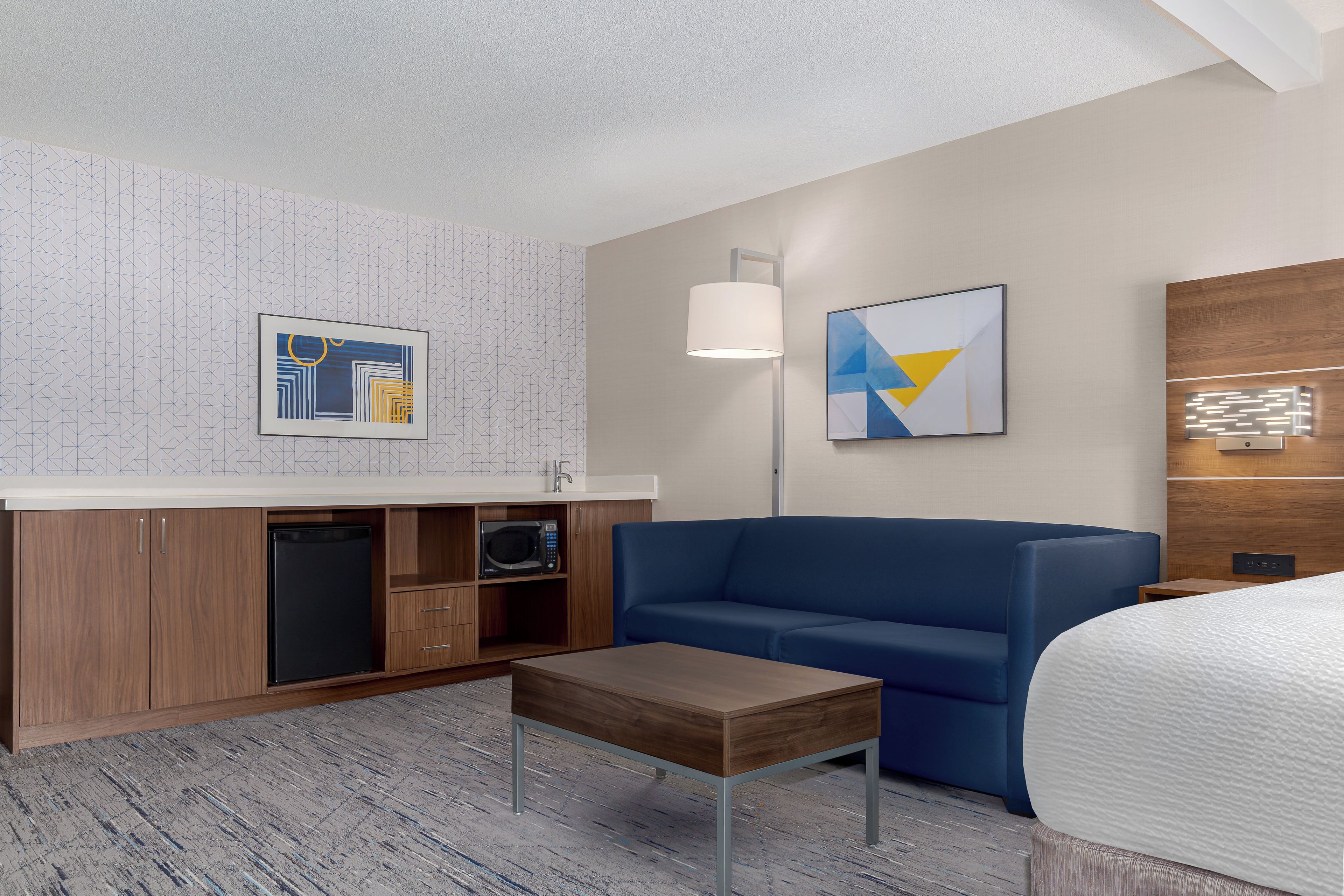 A hotel room with a blue sofa, a small wooden table, a microwave, and a cabinet with a sink.
