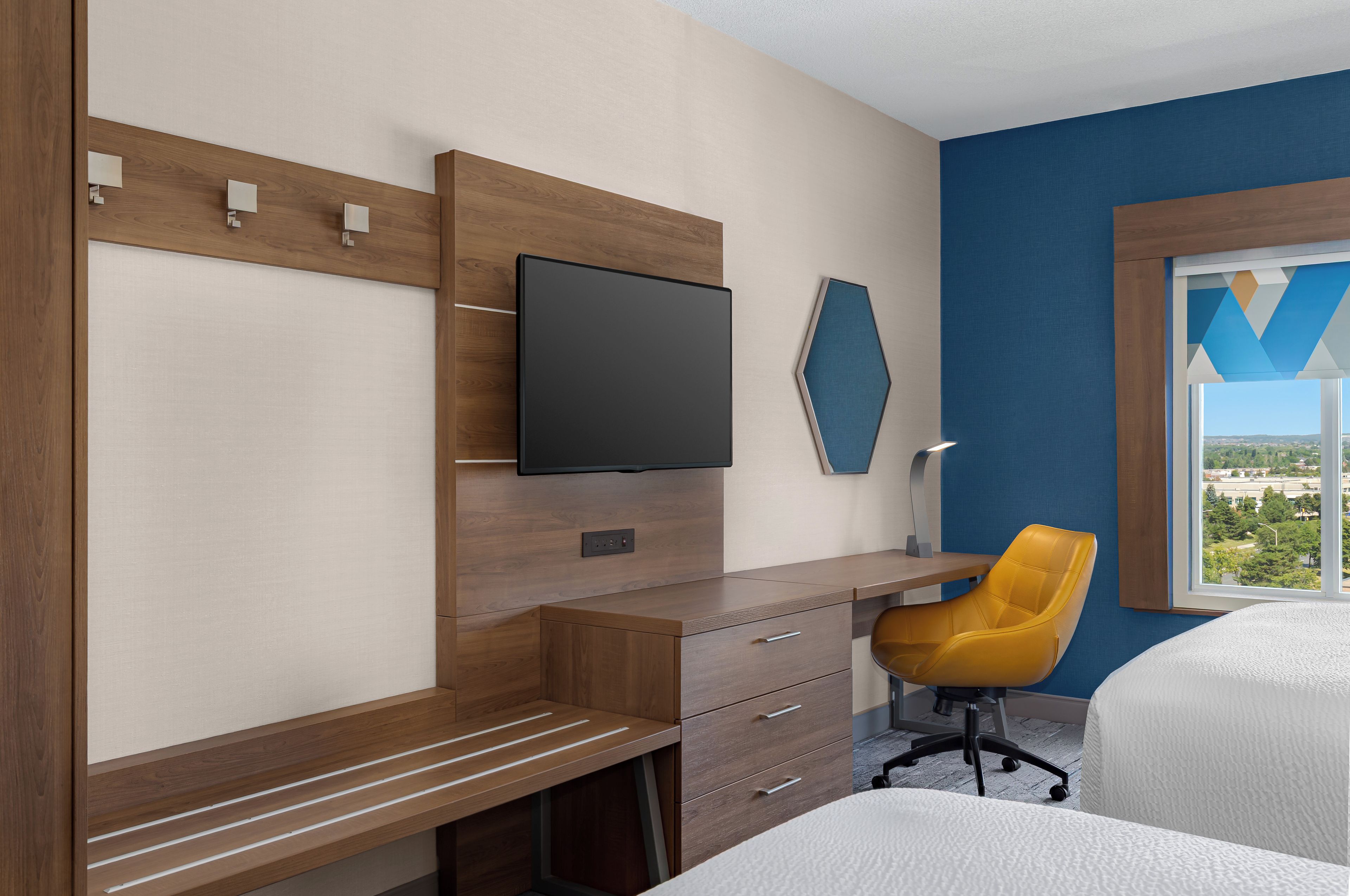 A hotel room with a wooden desk, a flat-screen TV, a yellow chair, and a bed.
