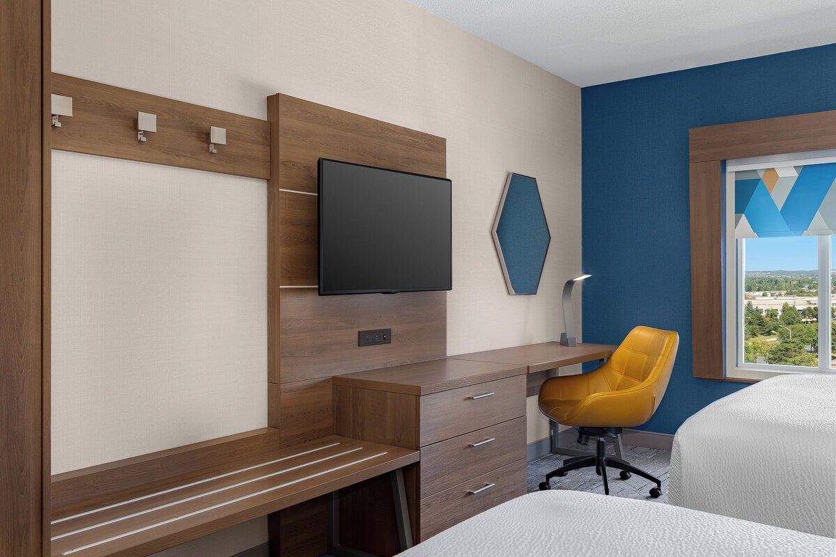 A hotel room with a wooden desk, a flat-screen TV, a yellow chair, and a bed.