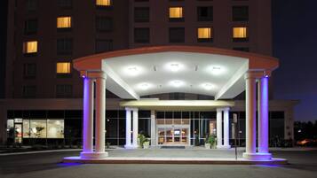 A hotel entrance with a covered waiting area, columns, and a building with lit windows in the background.