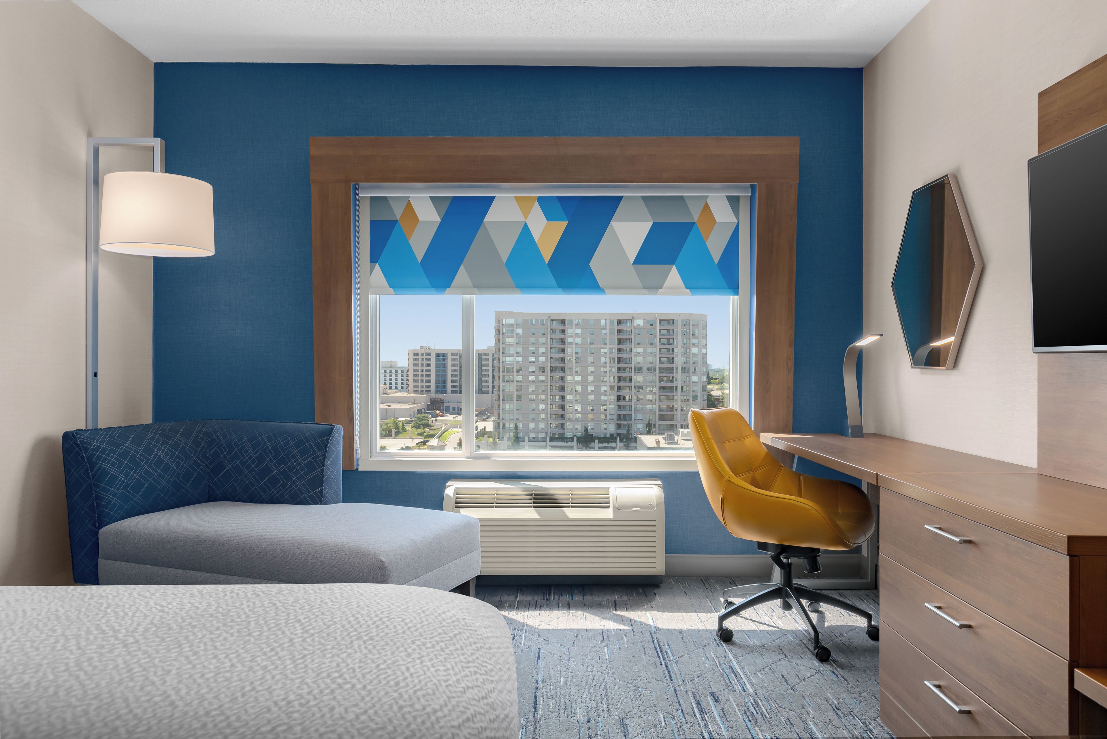 A hotel room with a large window, a desk, a chair, a bed, and a dresser.