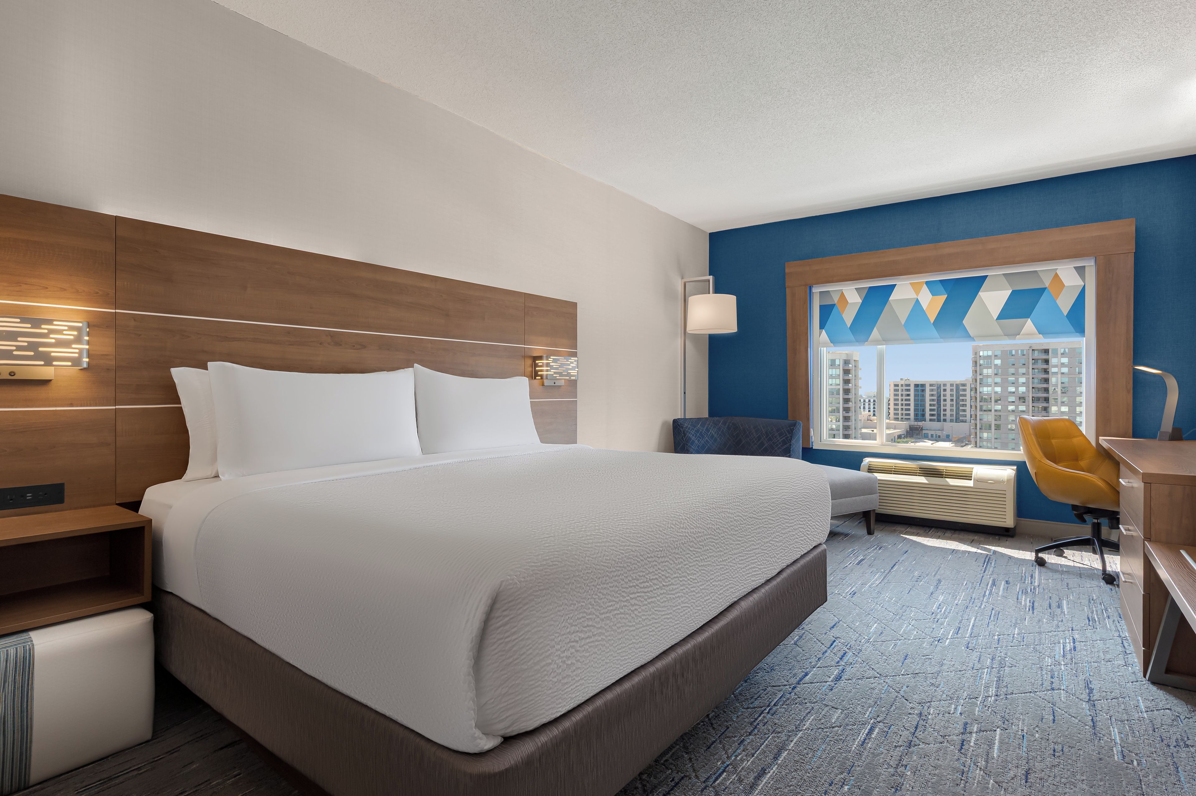 A hotel room with a large bed, a desk with a chair, a window with a city view, and a blue accent wall.