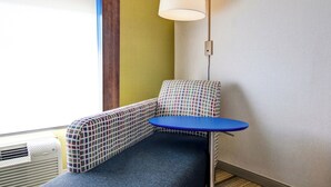 A corner room with a blue table, a patterned sofa, a white lamp, and a window with blinds. - Holiday Inn Express Hotel & Suites Toronto - Markham by IHG (Richmond Hill)