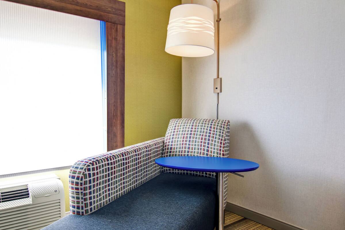 Une pièce d’angle avec une table bleue, un canapé à motifs, une lampe blanche et une fenêtre avec des stores.