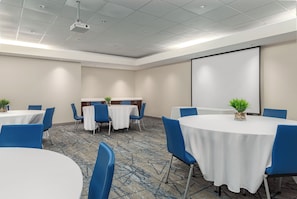 Une salle de conférence avec des tables rondes, des chaises bleues et un écran de projection.