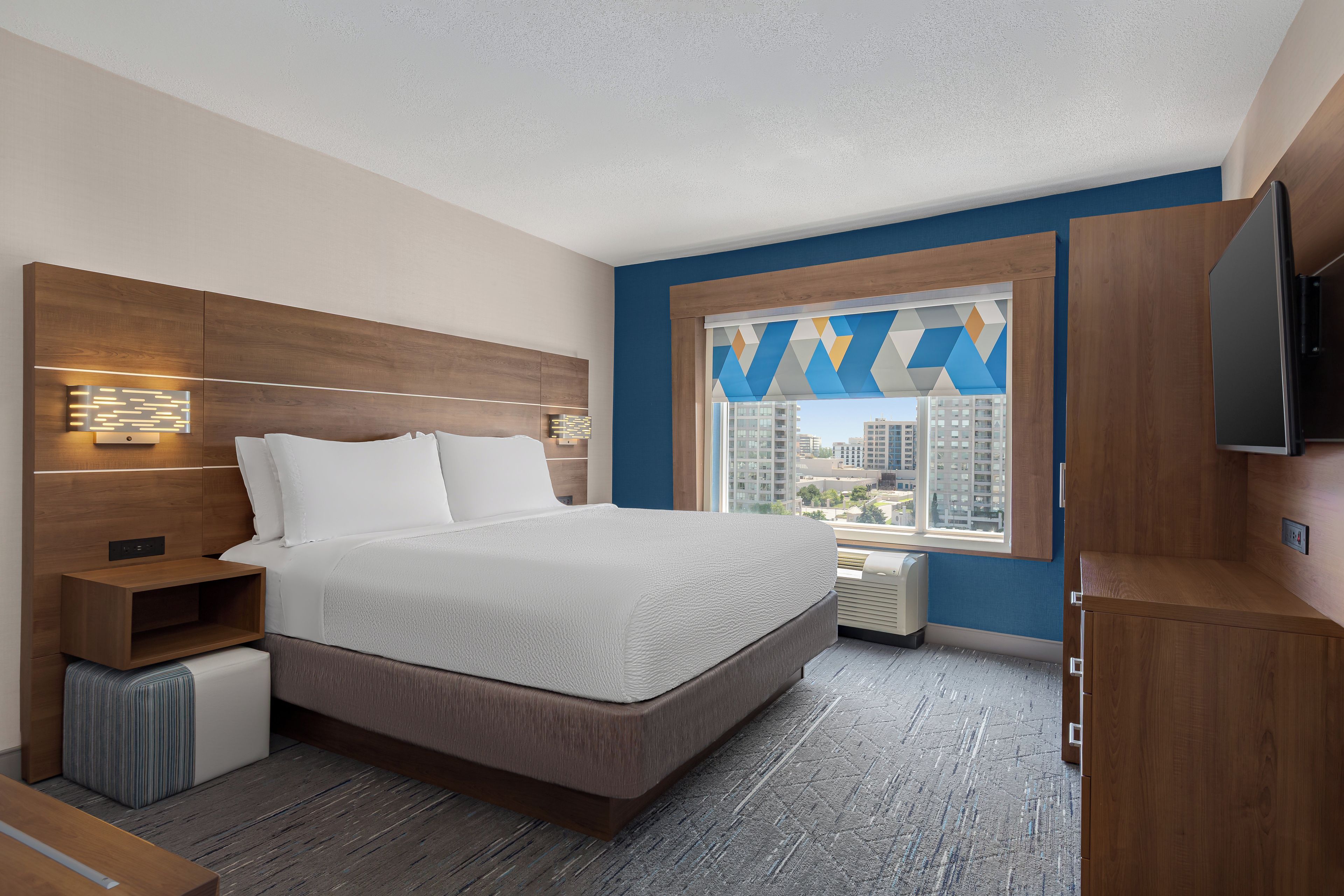 Een hotelkamer met een bed, een televisie, een raam met uitzicht op gebouwen en een houten hoofdeinde.
