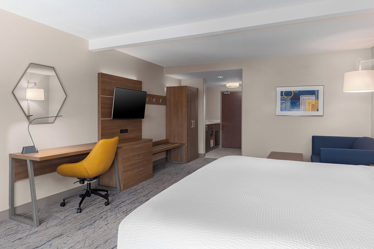 A hotel room with a bed, a desk with a chair, a television, a door, and a painting on the wall.