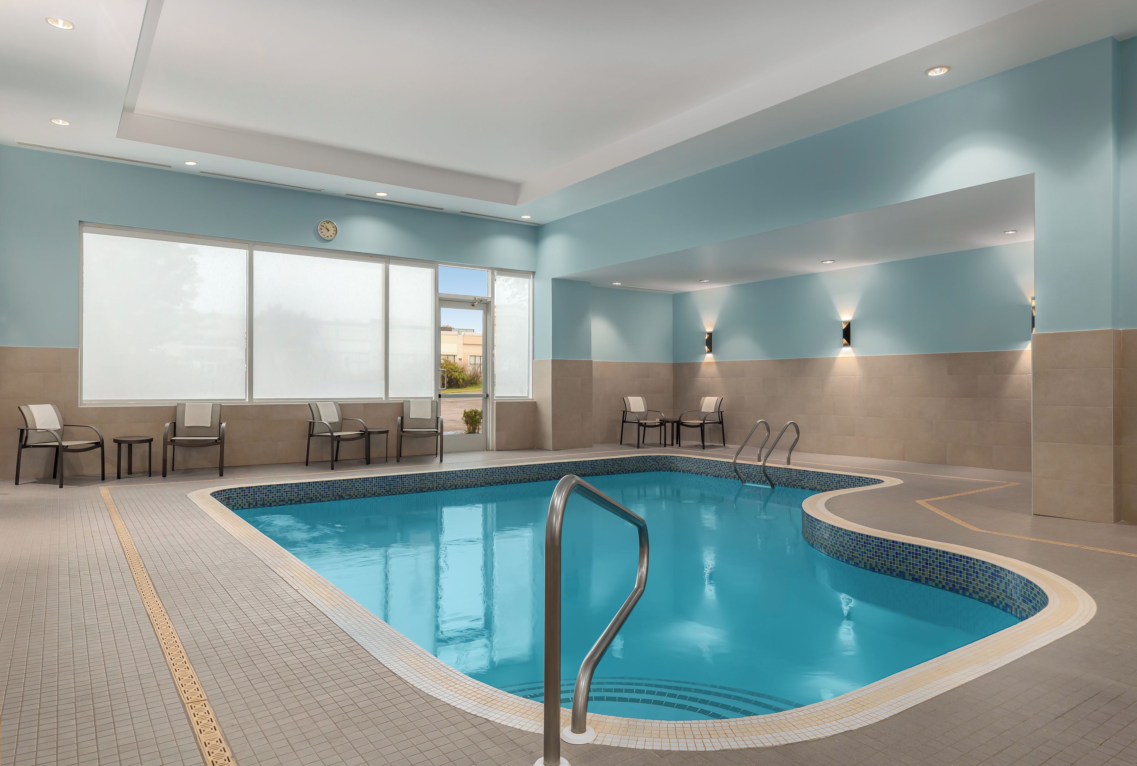 An indoor swimming pool with a tiled floor, a metal pool ladder, and chairs along the poolside.