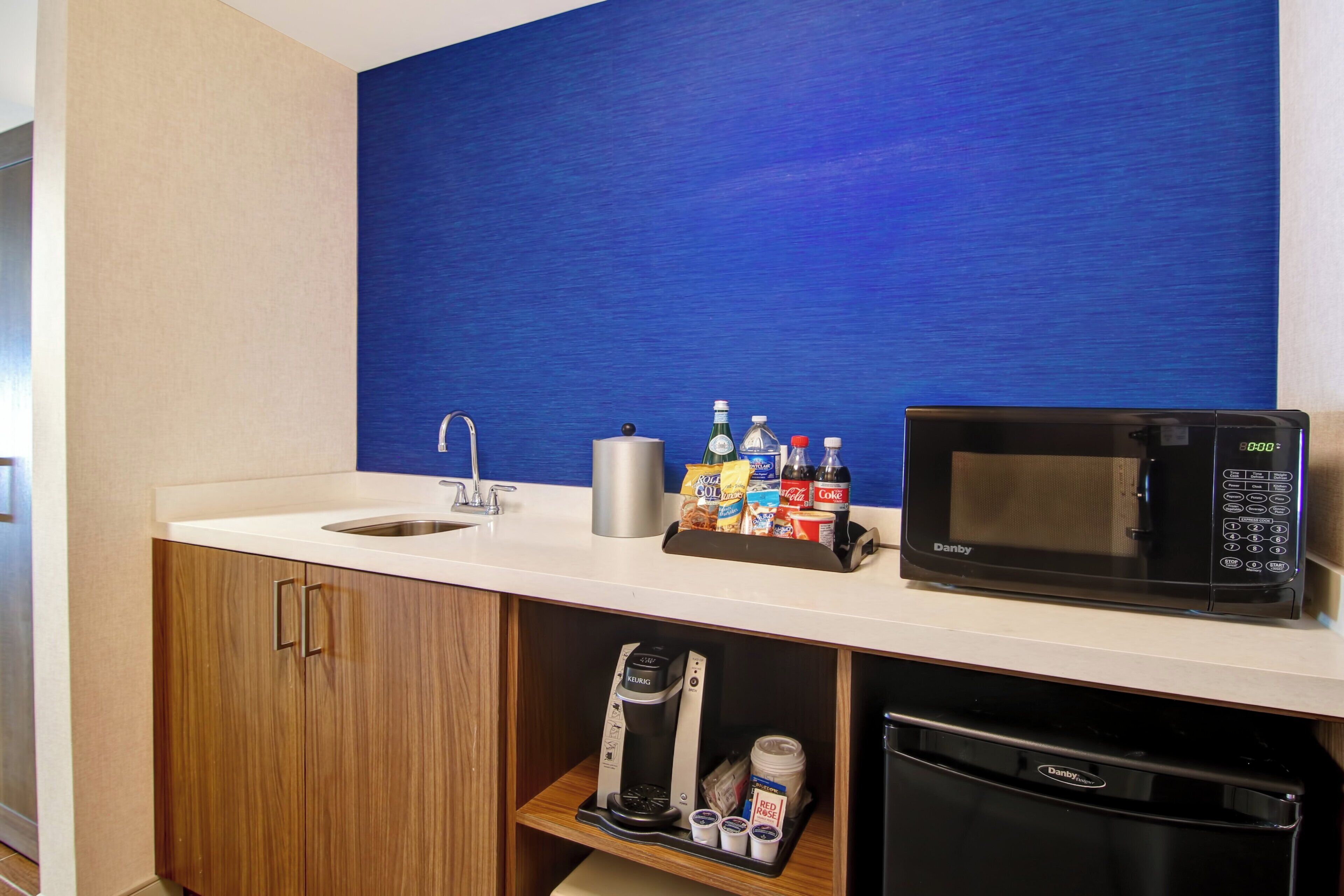 A kitchenette with a microwave, coffee maker, and a variety of food and drink items.