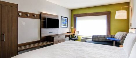 Une chambre d’hôtel avec un grand lit, un bureau avec une chaise, un téléviseur à écran plat, une fenêtre avec des stores et une lampe fixée au mur.