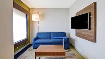 Una sala de estar moderna con un sofá azul, una mesa de centro de madera, una pequeña mesa redonda, una lámpara de pie y un televisor montado en la pared.