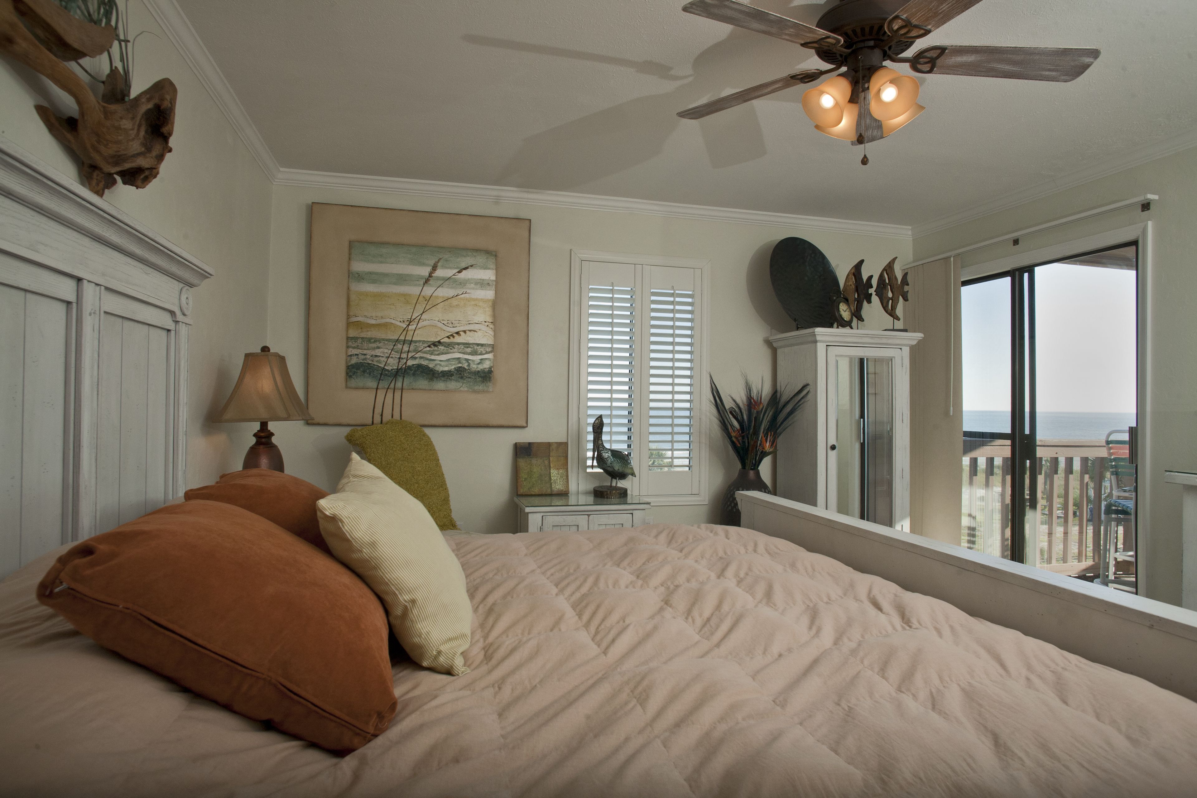 A bedroom with a large bed, a ceiling fan, a window with a view of the beach, and a nightstand with a lamp.