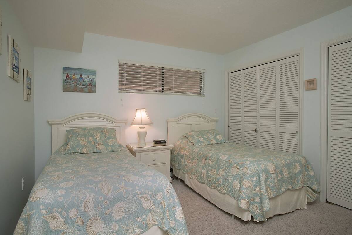A bedroom with two beds, a nightstand with a lamp, a painting on the wall, and a closet with louvered doors.