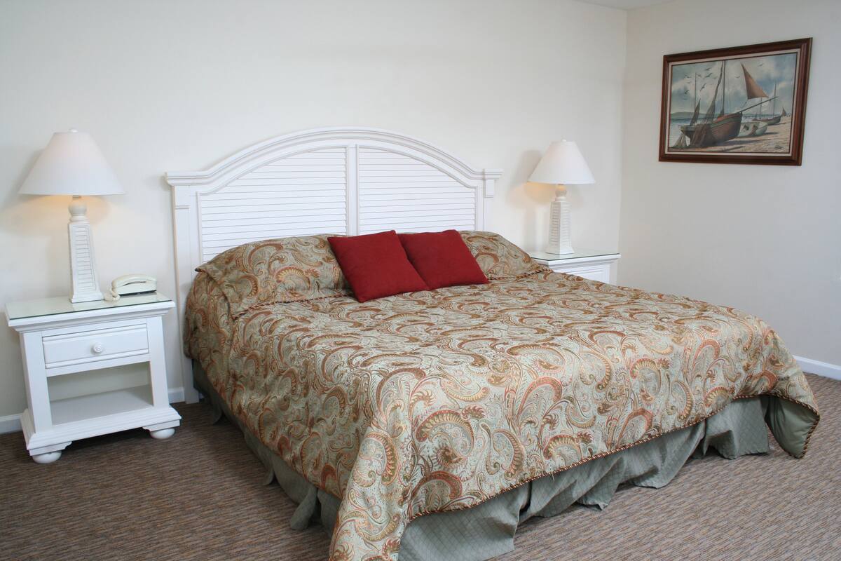 A neatly made bed with a patterned bedspread and two red pillows, flanked by white nightstands with lamps and a telephone.