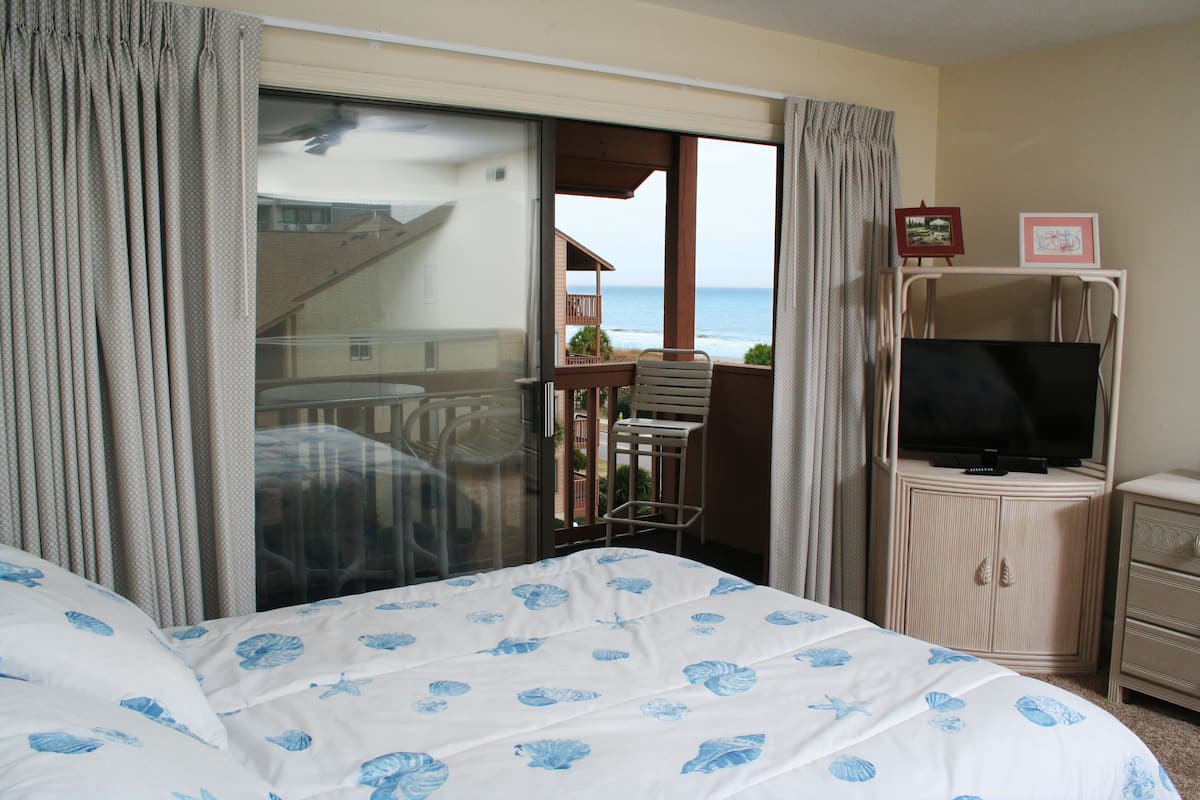 A hotel room with a bed, a television, a small table, and a view of the ocean through a sliding door.