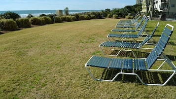 A row of metal sun loungers on a grassy lawn with a view of the ocean and a building in the background.