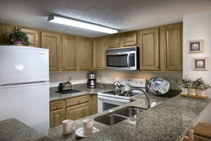 Una cocina con armarios de madera, un microondas de acero inoxidable, un refrigerador blanco, encimera de granito, un lavamanos y una cafetera.