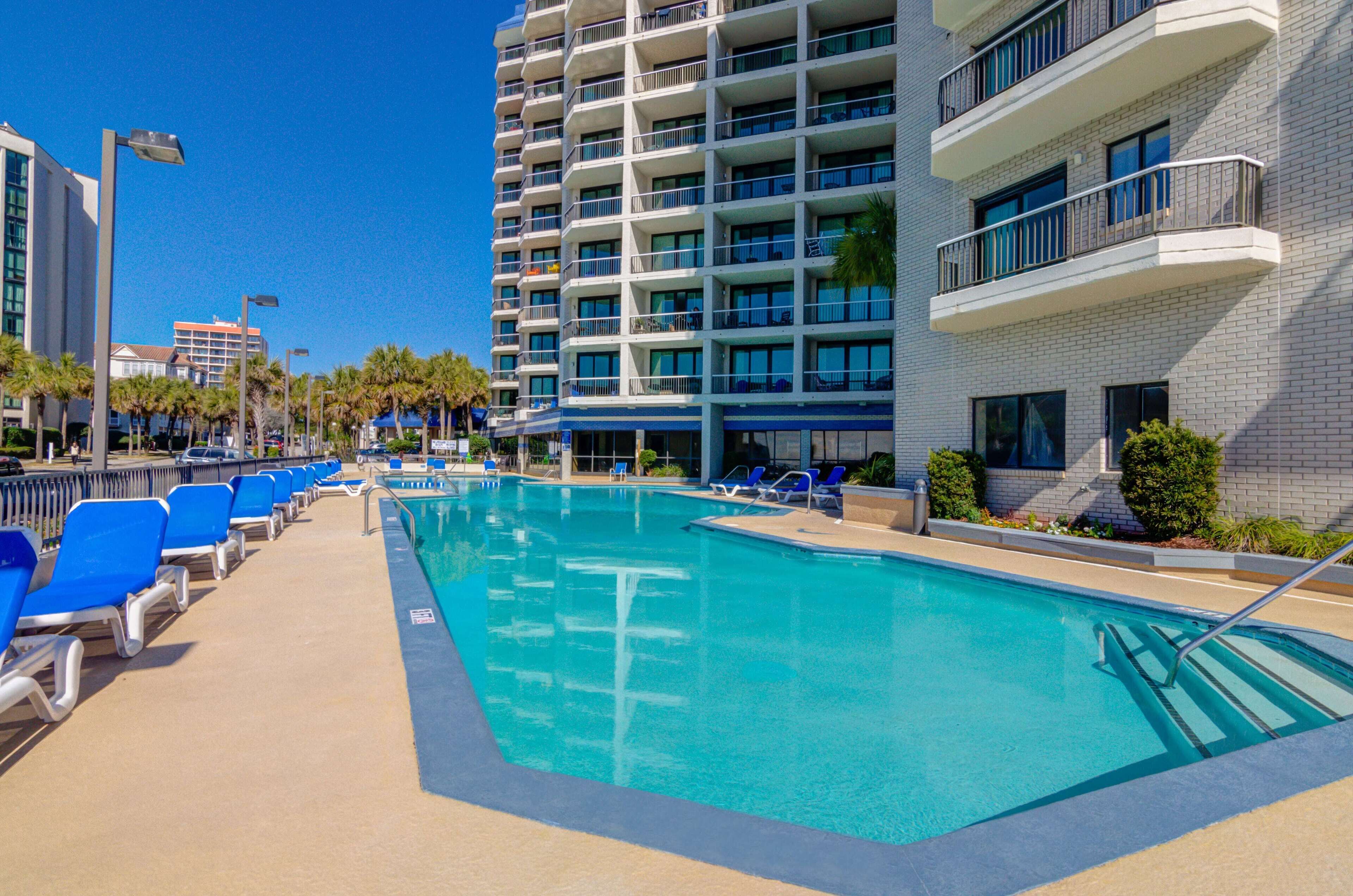 Zona de piscina de hotel con sillas reclinables azules, una piscina de color azul cristalino y un edificio de varios pisos con balcones.