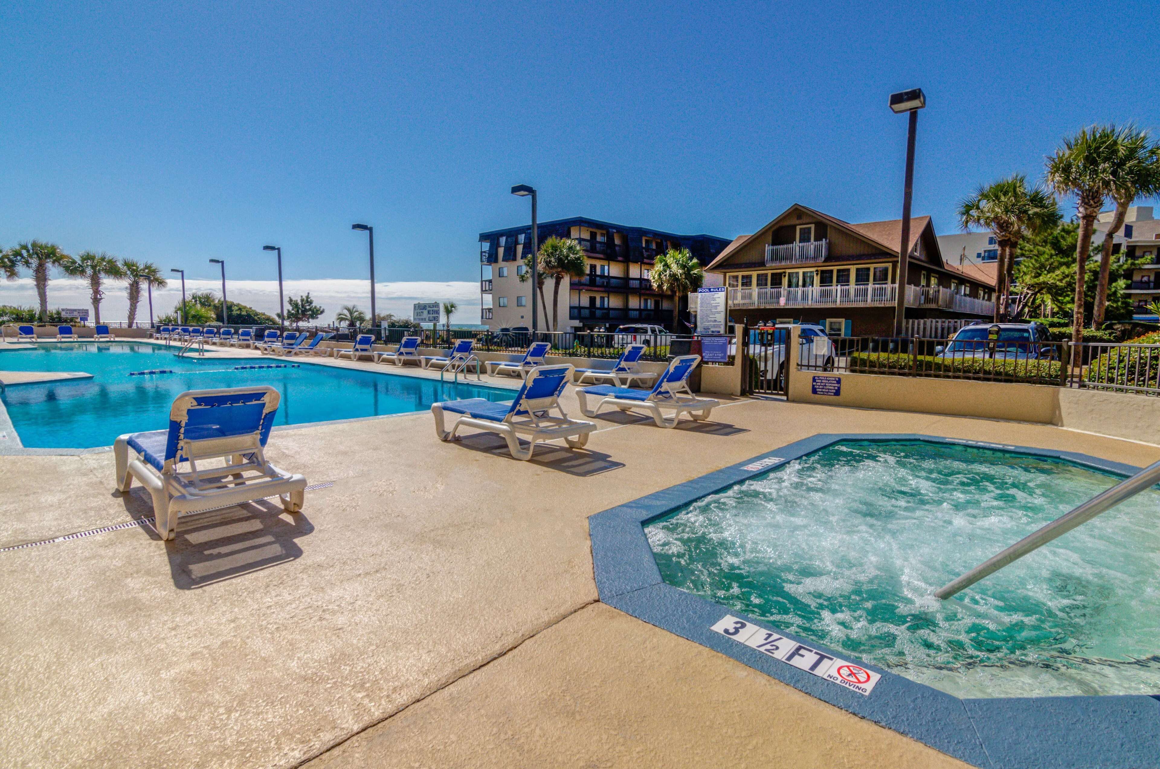 A swimming pool area with lounge chairs and a hot tub, surrounded by buildings and palm trees.