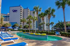 A pool area with lounge chairs, palm trees, and a hotel building with balconies. - Carolina Winds (Myrtle Beach)