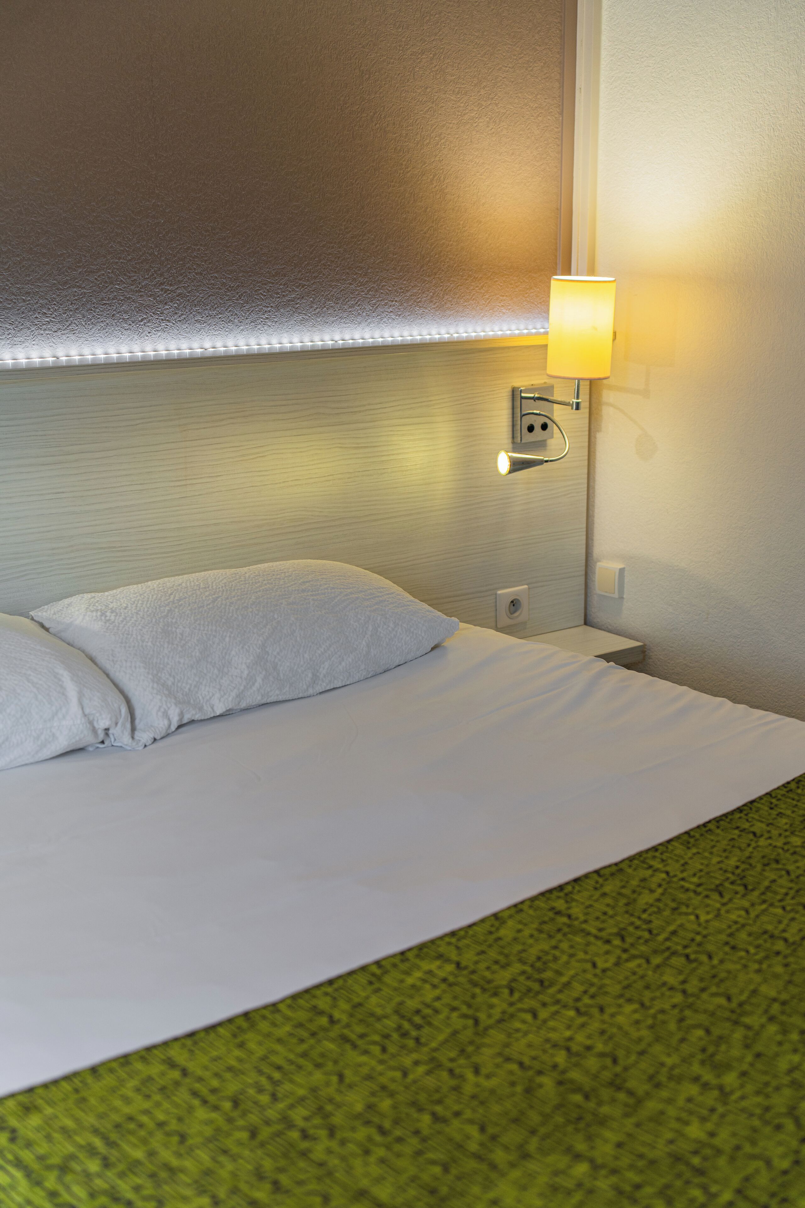A hotel room with a neatly made bed, a wall-mounted lamp, and a textured wall.