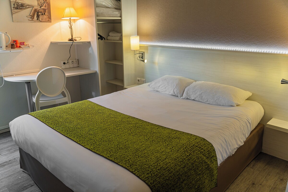 Een opgemaakt bed met een groene plaid, een nachtkastje, een stoel en een lamp.