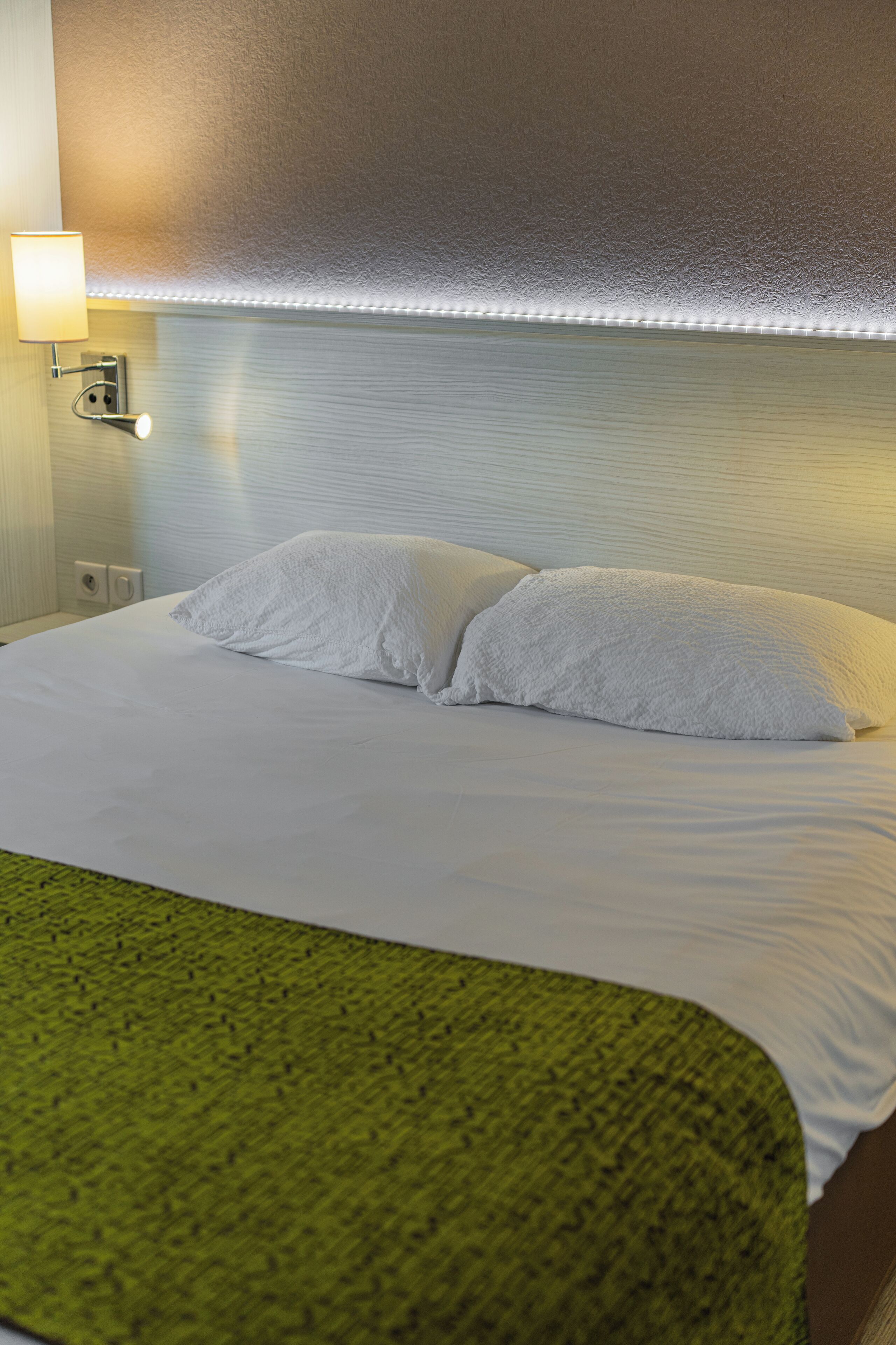 A neatly made bed with white linens and a green throw blanket, a wall-mounted lamp, and a textured wall.