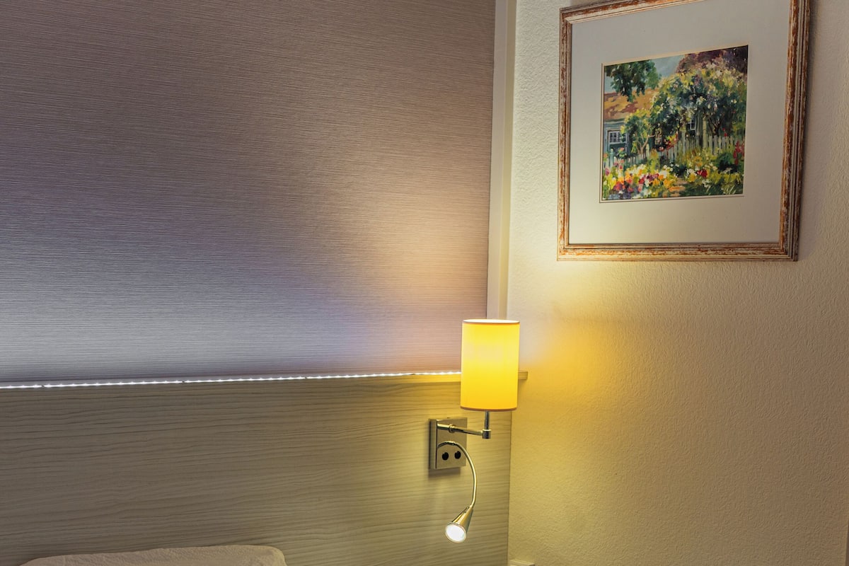 Una habitación de hotel con una cama bien hecha, una manta roja, una lámpara de pared y un cuadro en la pared.