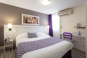 Une chambre d’hôtel comprenant un lit, une table de chevet, une chaise, un bureau, un appareil de climatisation, un tableau et une salle de bain avec un rideau de douche.