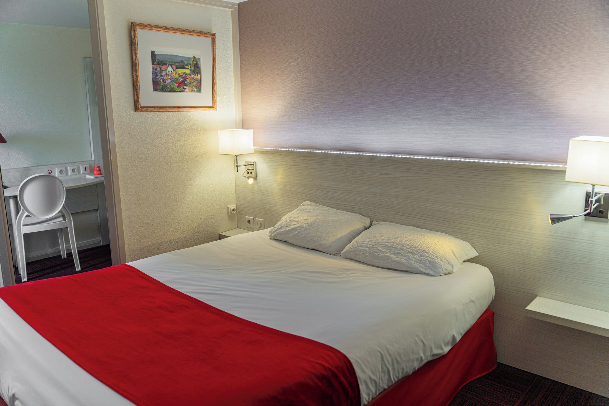 A hotel room with a bed, a desk, a chair, and a wall-mounted lamp.
