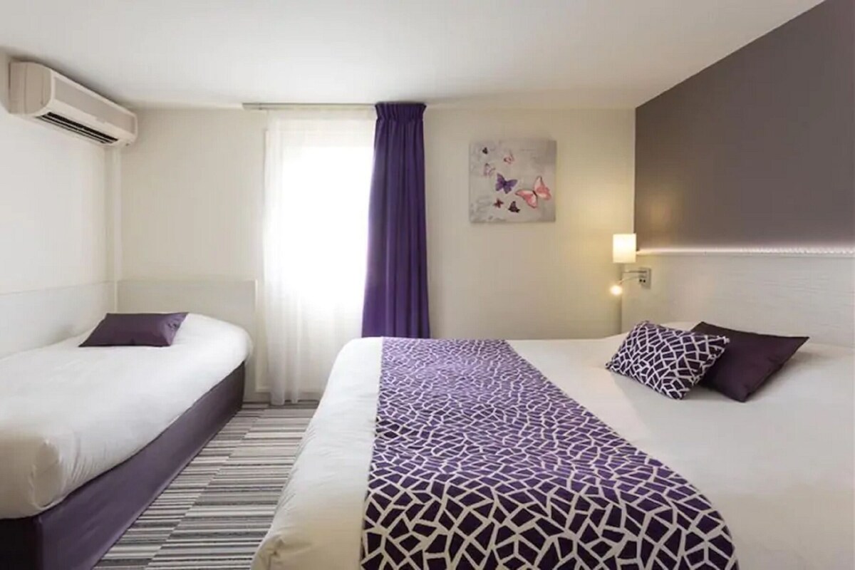Hotelkamer met twee bedden, een raam met paars gordijn, een wandairconditioner en een kunstwerk aan de muur.