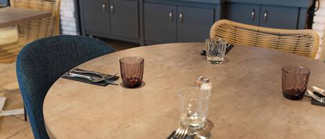 Una mesa redonda con mantel beige, dispuesta con cubiertos y vasos, en una habitación con sillas de mimbre y un armario azul al fondo.
