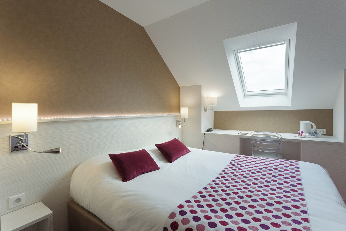 Une chambre d’hôtel comprenant un lit, deux oreillers, une table de chevet, une lampe, un bureau avec une chaise, une fenêtre et un miroir.