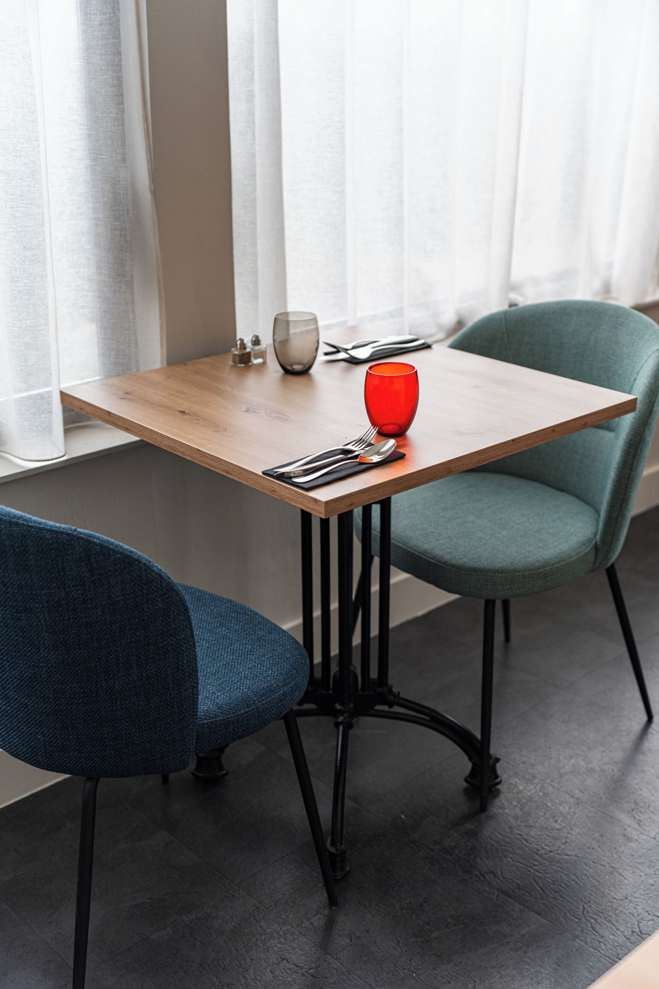 A table with a red vase, a glass, and cutlery, flanked by two chairs, one blue and one teal, in a room with a window and curtains.