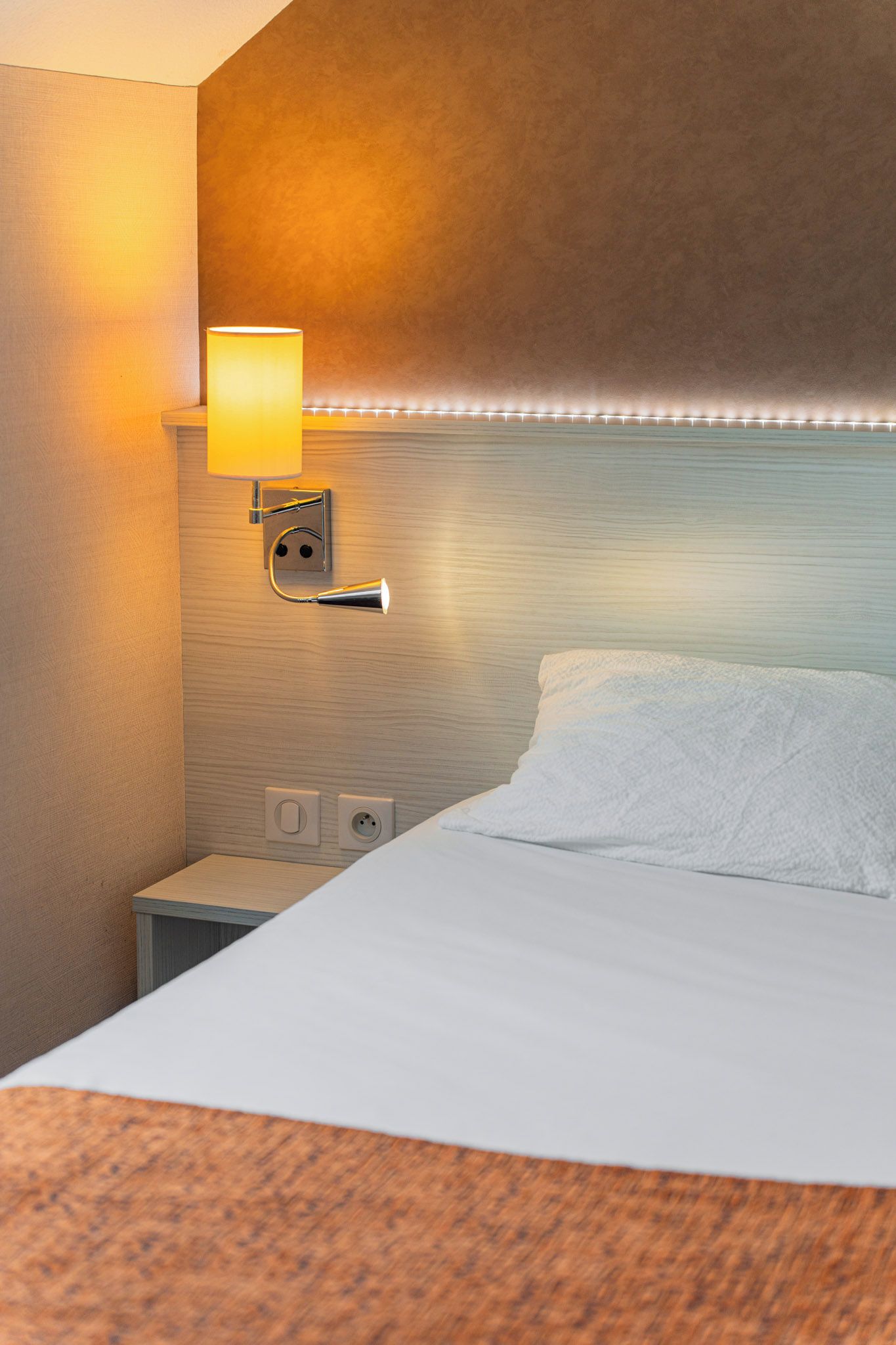 A hotel room with a bed, a nightstand, a wall-mounted lamp, and a power outlet.