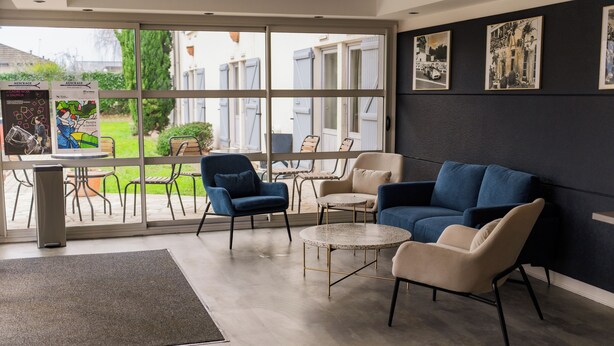Een moderne zithoek met een blauwe en beige bank, een kleine ronde tafel en een bijzettafeltje. Grote ramen bieden uitzicht op het exterieur.