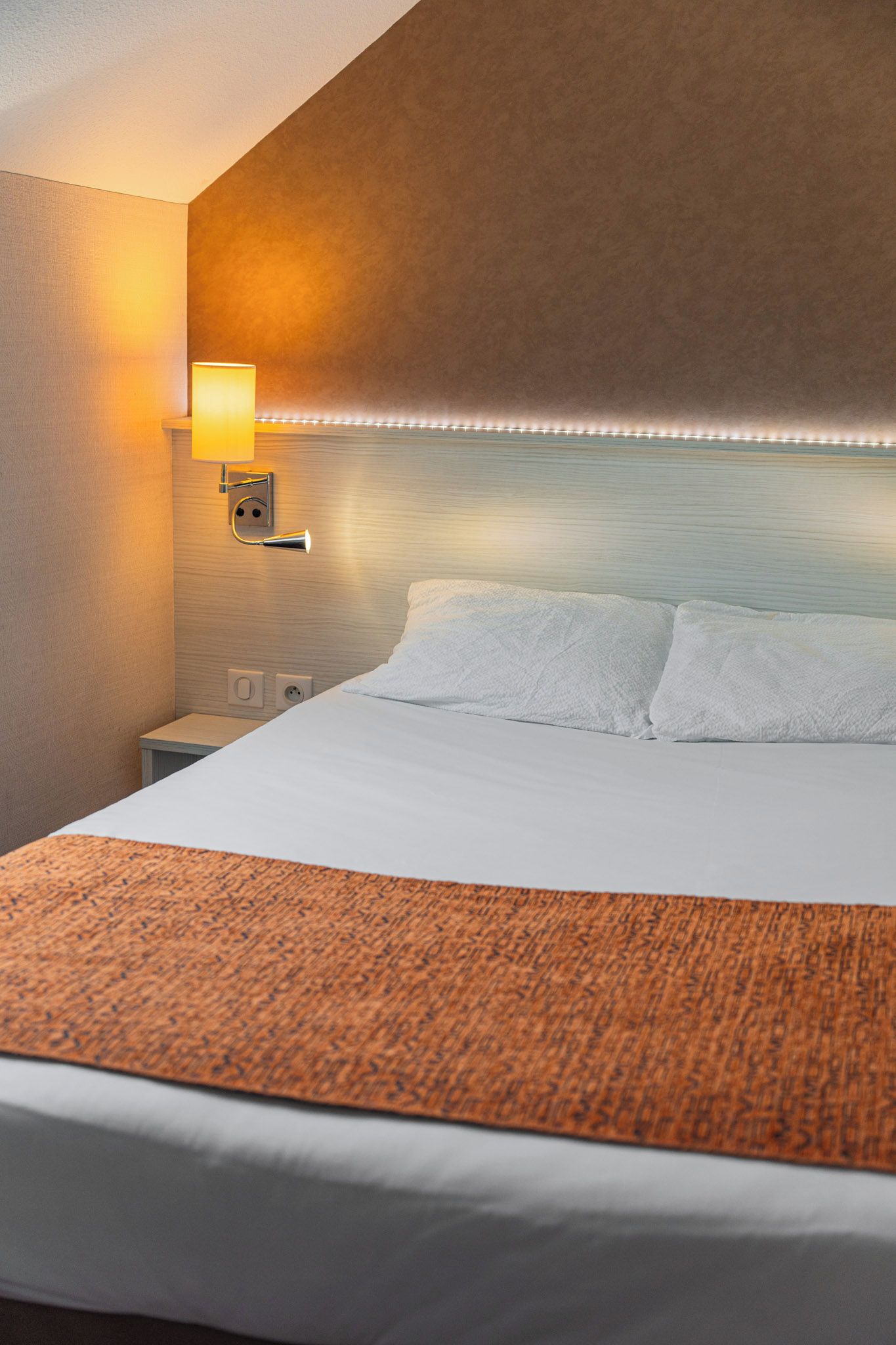 Una habitación de hotel con una cama, una mesita de noche, una lámpara de pared y una alfombra naranja con textura.
