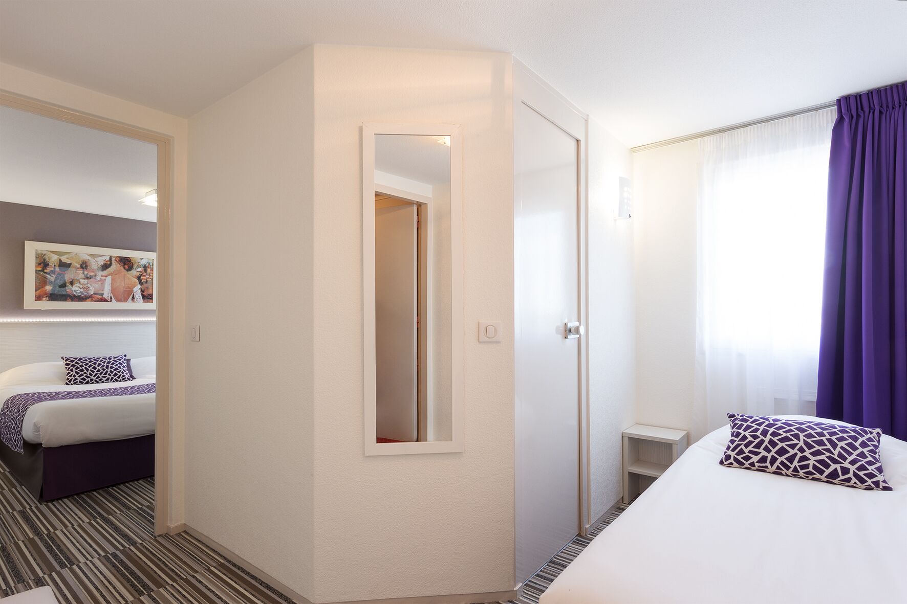 A hotel room with a bed, a bedside table, a mirror, a door, and a window with curtains.