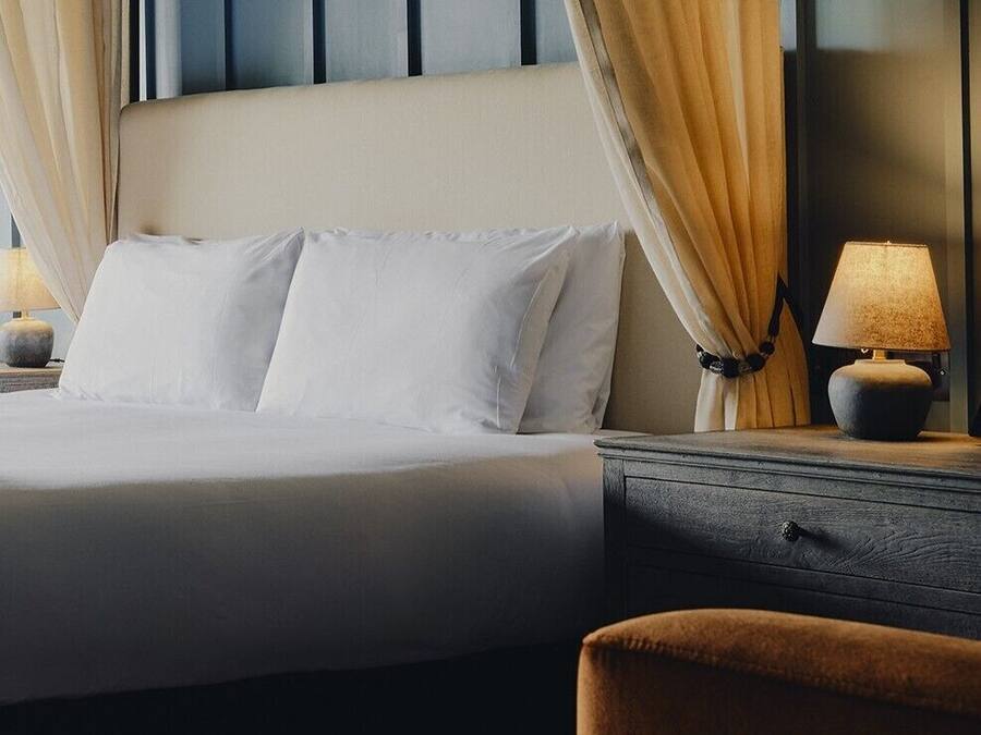 A neatly made bed with white linens, a bedside table with a lamp, and a chair.