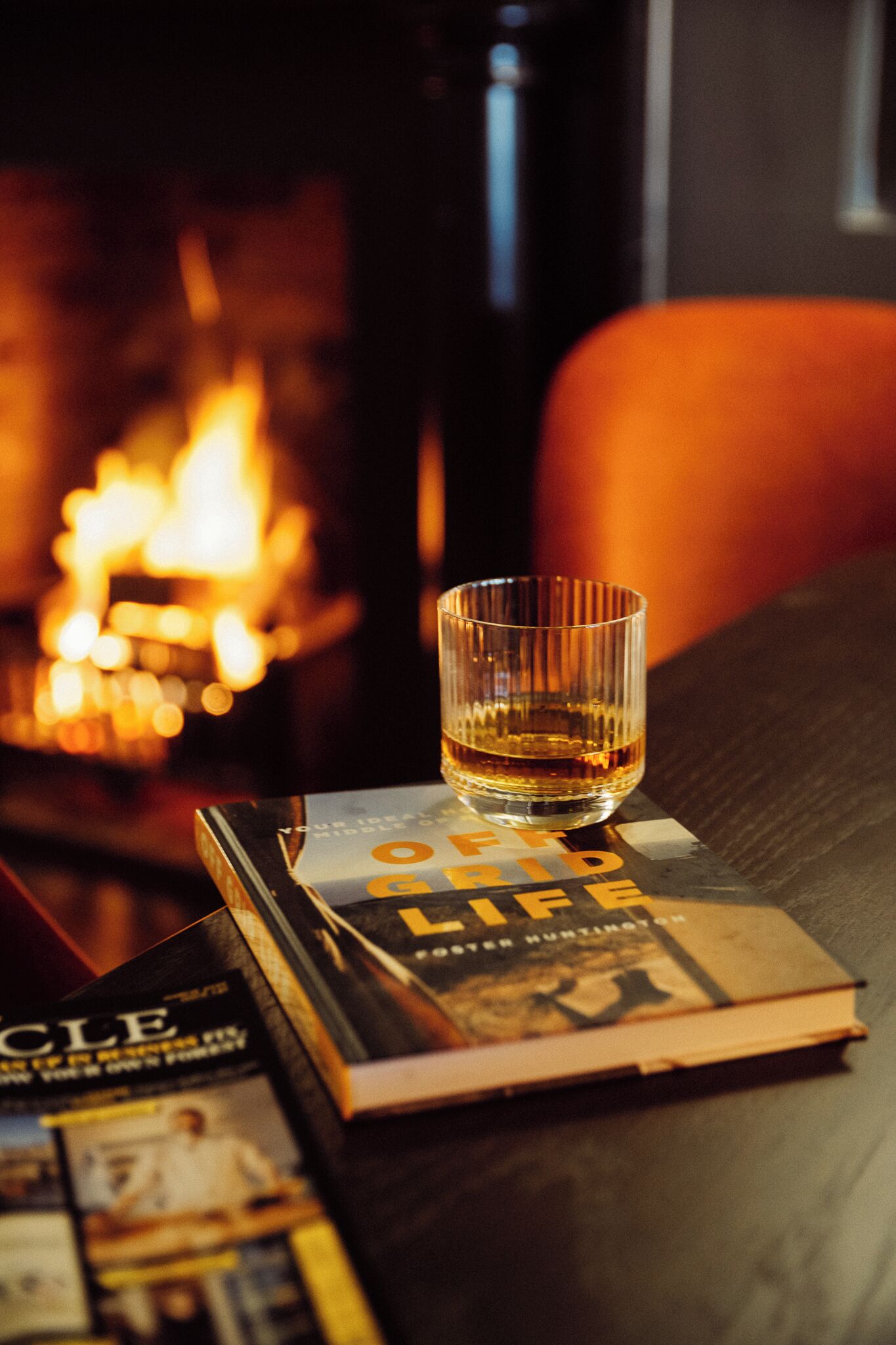 A glass of whiskey on a table with books and a fireplace in the background.