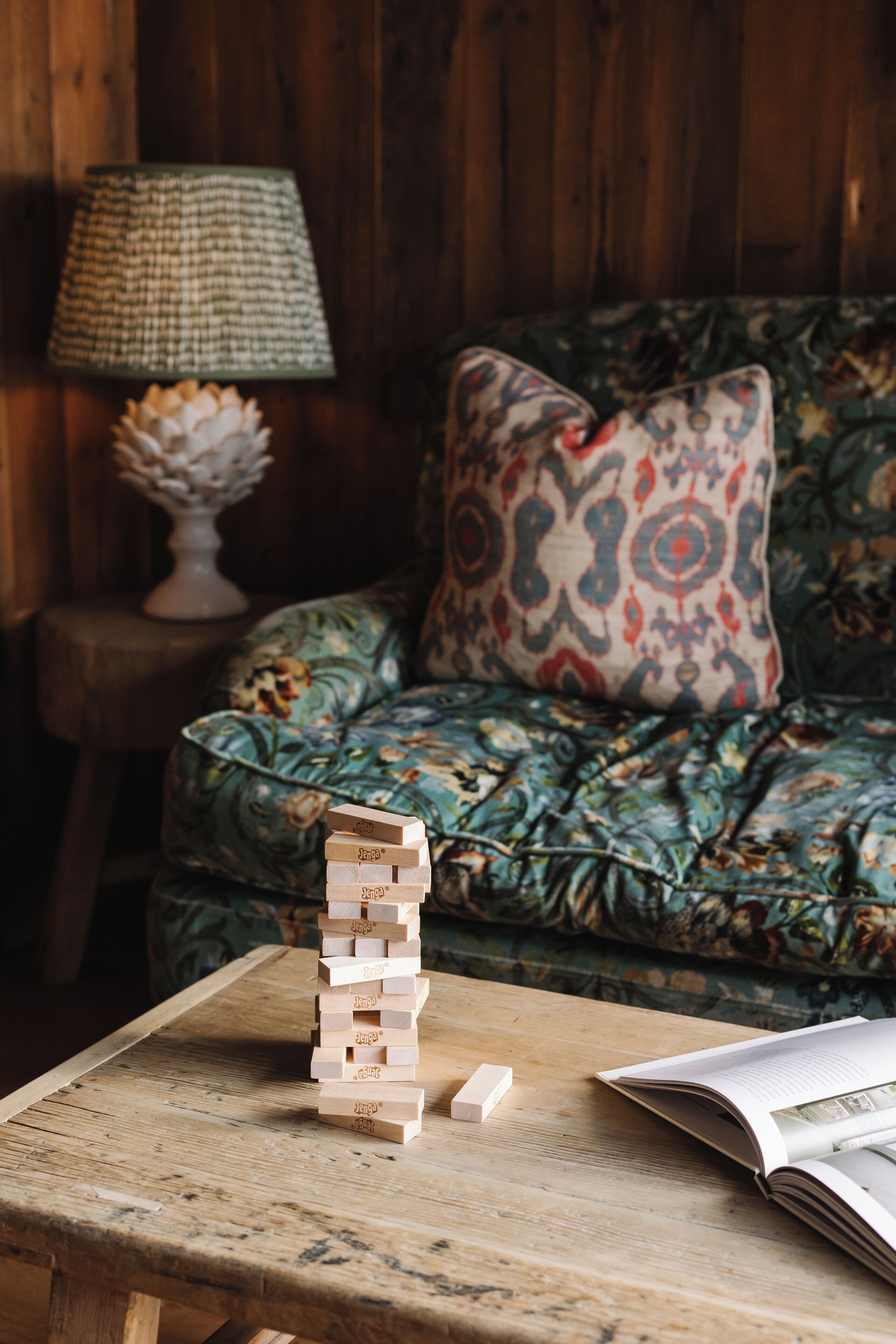 A wooden table with a stack of wooden blocks, a lamp, a patterned cushion, and an open book.